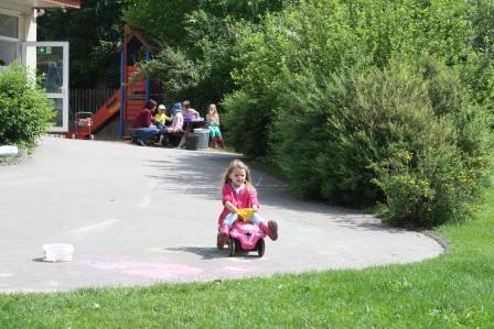 Ein Mädchen fährt auf einem Bobbycar über das Außengelände. Im Hintergrund sitzen weitere Kinder an einem Tisch