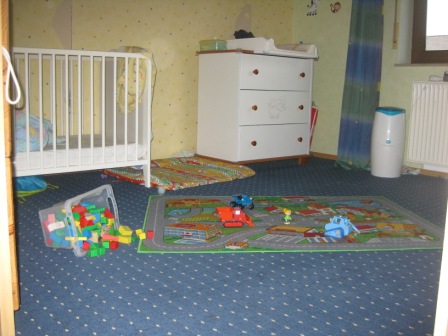 Im hell eingerichteten Kinderzimmer findet sich ein Kinderbett und eine Wickelkommode. Ebenso finden sich bunte Spielteppiche und Spielzeuge
