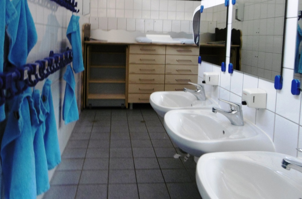 Der Waschbereich in der Kita. Hier sind niedrig angebrachte Waschbecken zu sehen. Im Hintergrund steht ein Wickeltisch. An der linken Wand sind Haken mit Handtüchern für jedes Kind angebracht.