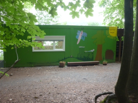 Der Waldcontainer von Außen: Ein grüner Container, in dem sich die Kinder bei schlechtem Wetter aufhalten können. Ein Schmetterling und eine Sonne sind außen aufgemalt.
