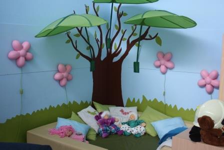 Das "Wohnzimmer" der Kita Niederscheld bietet Raum für das Freispiel. Hier zu sehen eine Puppenecke. an die Wand ist ein Baum gemalt, dessen Zweige aus kleinen Sonnensegeln in Blattform, schützend über den Spielbereich ragen. Weiterhin sind an den Wänden Lampen in Blumenform angebracht