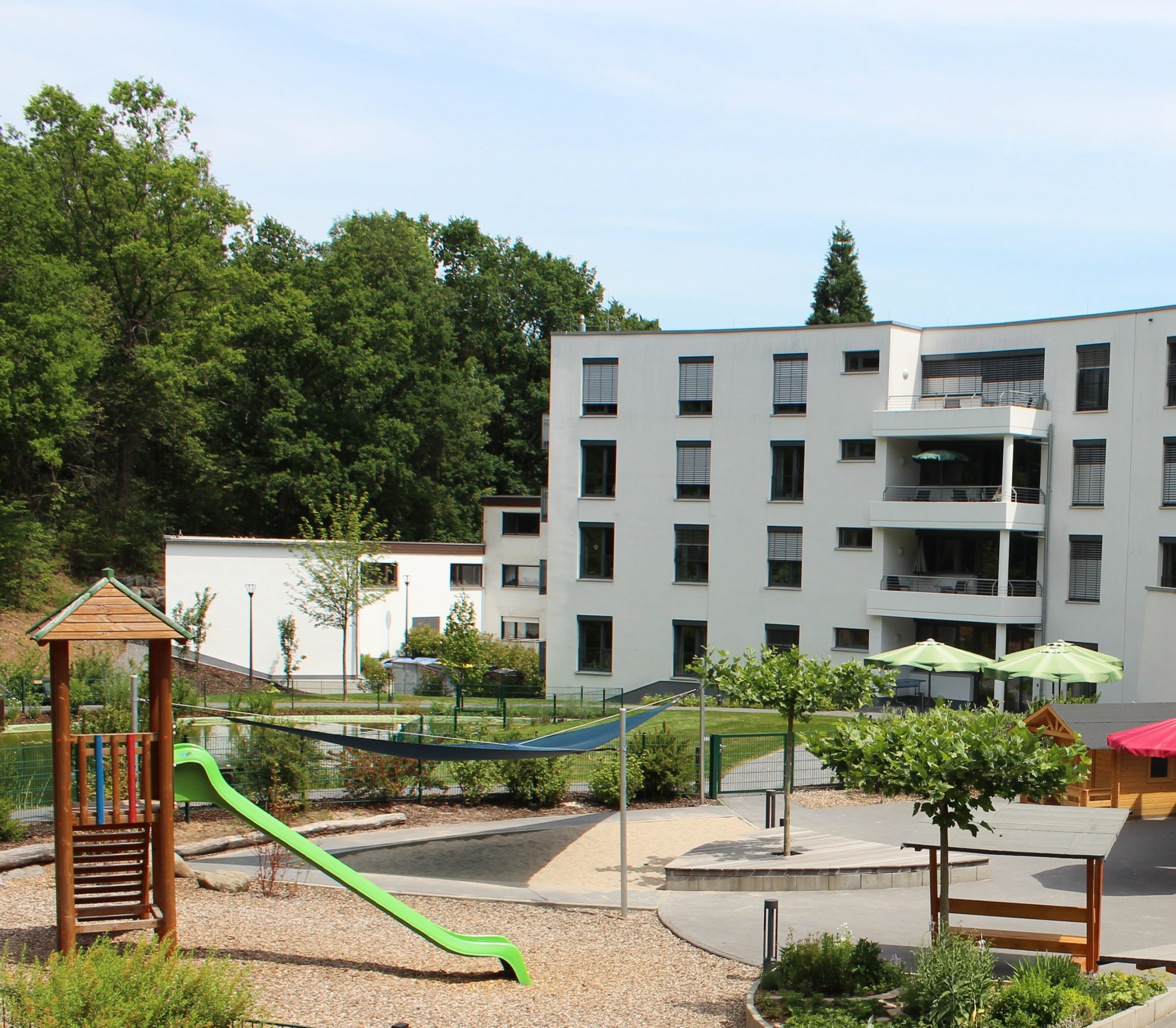 Heller Spielplatz mit Gebäude im Hintergrund: hellgrüne Rutsche, großer Sandbereich mit Sonnensegel, Außenbepflanzung grüne Sträucher sowie vereinzelte junge Platanenbäumen auf dem Gelände.