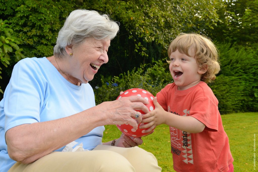 Lachende Seniorin, im Spiel mit Kita-Kind das lachend und mit Begeisterung roten Ball mit weißen Punkten an die Seniorin reicht
