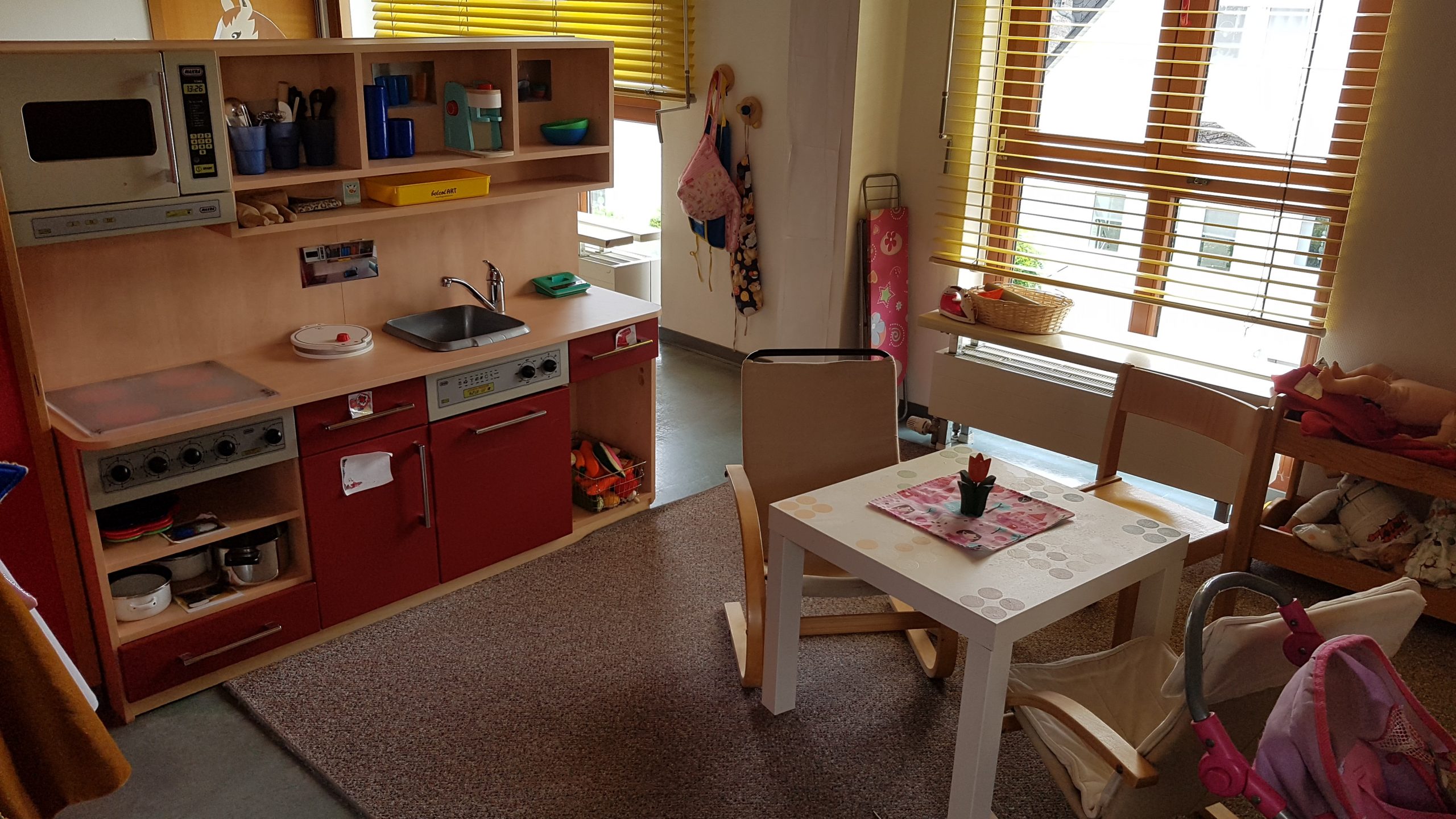 Eine Kinderküche mit Sitzbereich laden zum Freispiel ein. Die Schränke der Küche sind rot und es gibt eine Mikrowelle, einen Herd, eine Spüle und eine Spülmaschine, ganz wie in einer richtigen Küche. In der rechten Ecke daneben steht ein Kinderbügelbrett.