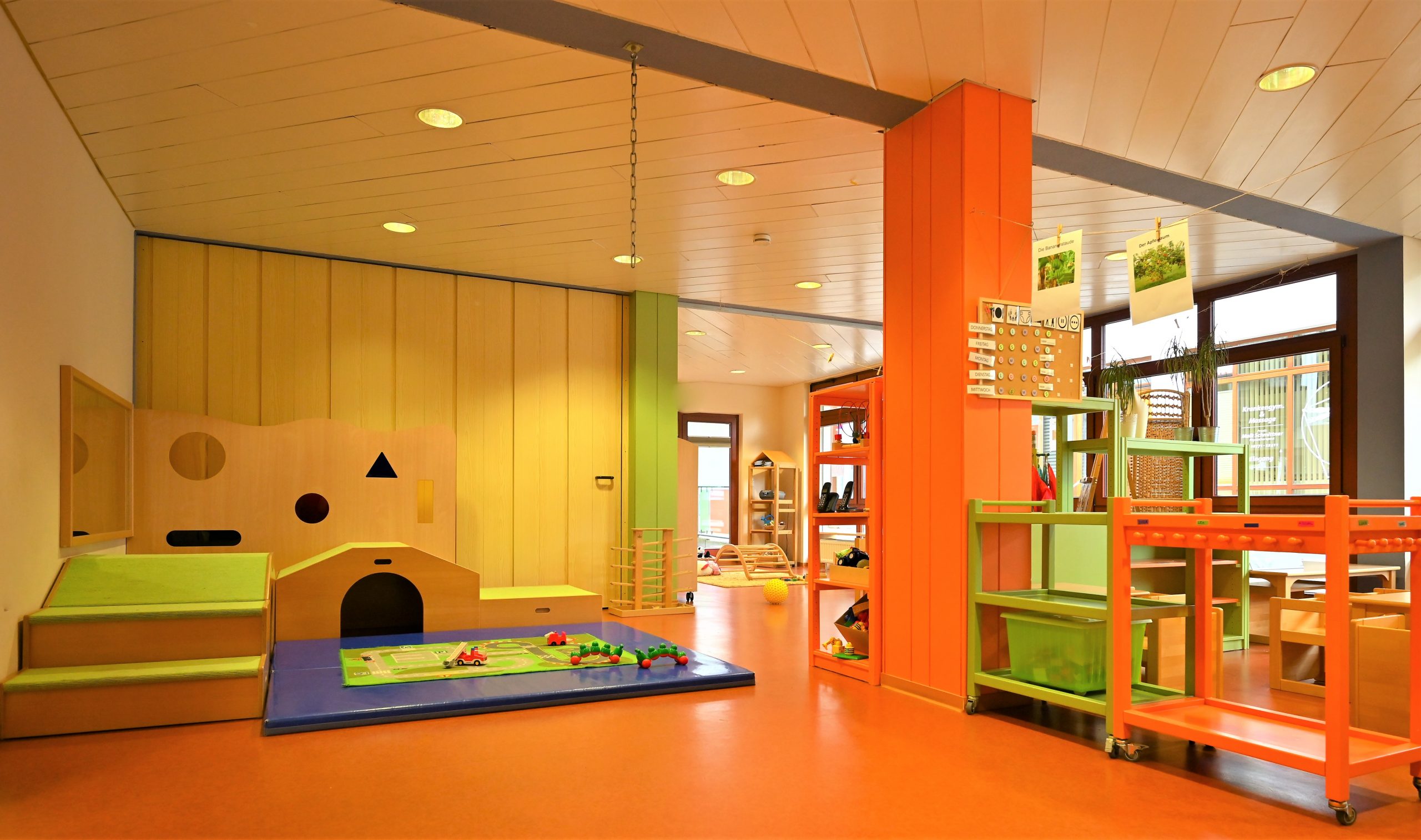 Der Krippenraum ist ein großer heller Raum mit einer hellen Holzdecke und orangefarbenem Boden. Die Möbel und Aufbewahrungen sind in grün und orange gehalten. Vor einem Spielgerät zum Klettern und Krabbeln, welches mit grünem Teppich bezogen ist, findet sich eine blaue Turnmatte.